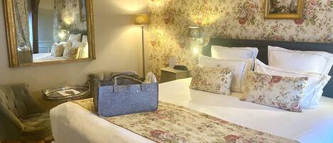 A hotel room with a large bed, floral wallpaper, a mirror, a nightstand, a lamp, and a chair.