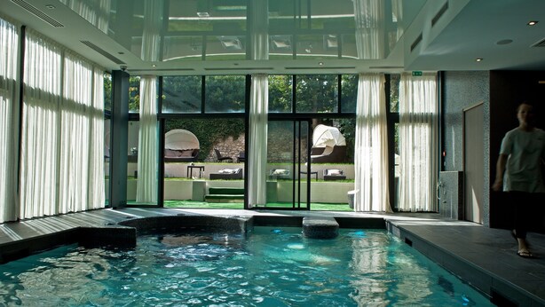 An indoor swimming pool with clear glass walls, a view of the outdoors, and a seating area with umbrellas.