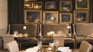 A dining area with a round table, chairs, and a chandelier. There are framed artworks on the wall behind the seating.