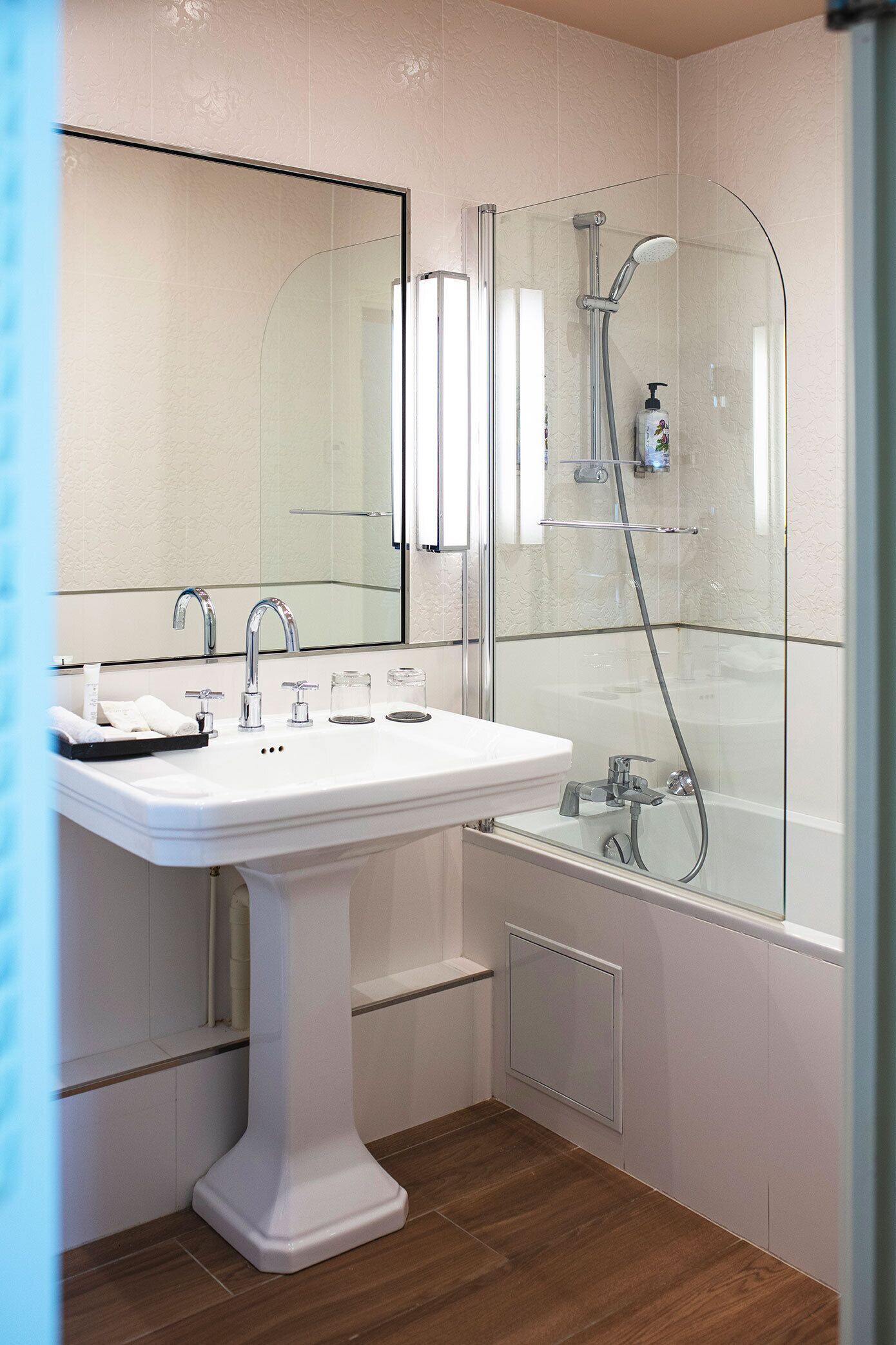 A bathroom with a white pedestal sink, a large mirror, a glass-enclosed shower, and a wooden floor.