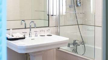 A bathroom with a white pedestal sink, a large mirror, a glass-enclosed shower, and a wooden floor.