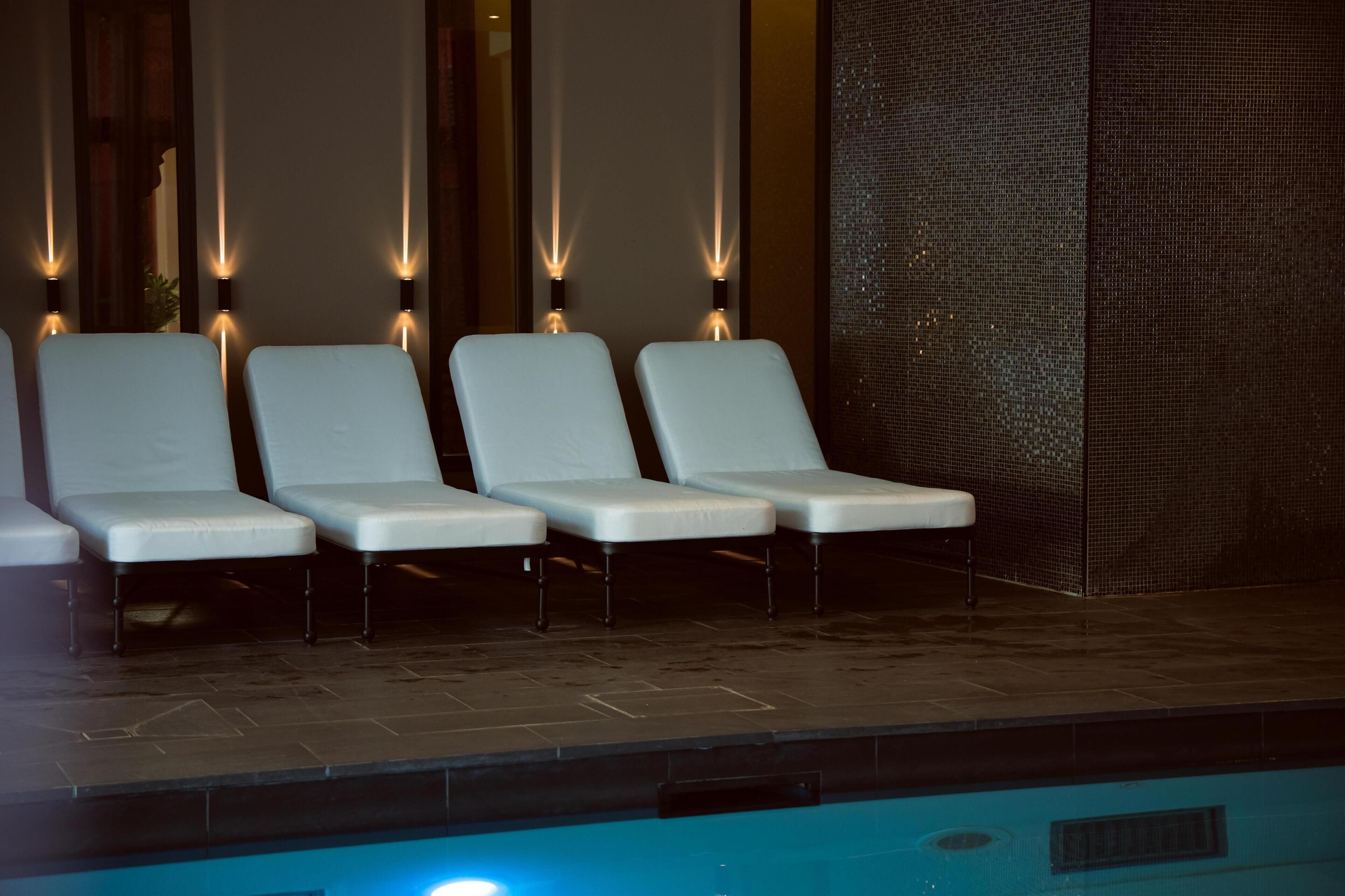A poolside area with four white lounge chairs, a tiled floor, and a decorative wall with vertical light fixtures.