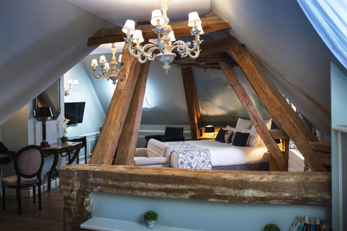 Um quarto com teto inclinado, vigas de madeira aparentes, um candelabro, uma cama com almofadas, uma mesinha e uma cadeira.