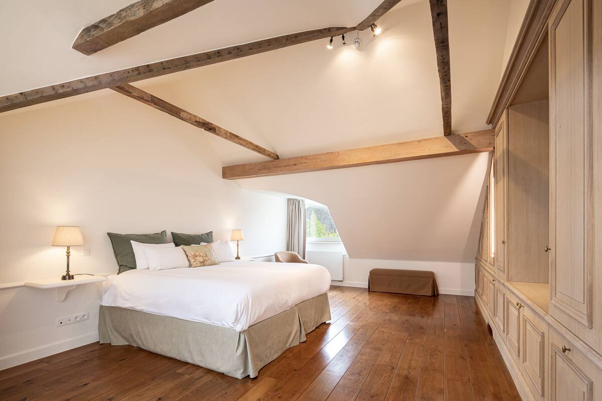 A bedroom with a wooden ceiling, a large bed, a nightstand with a lamp, a bench, and a wardrobe.