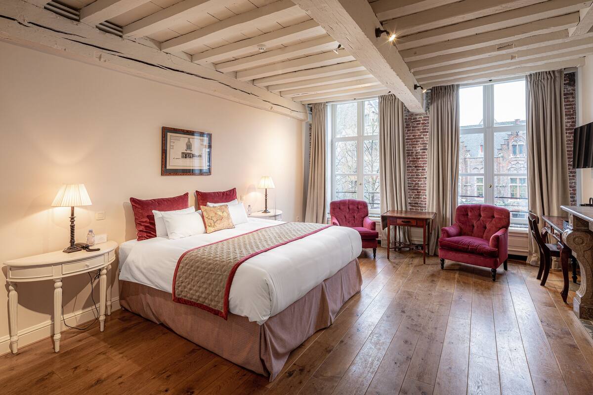 A bedroom with a large bed, a desk, a chair, and a window with curtains.