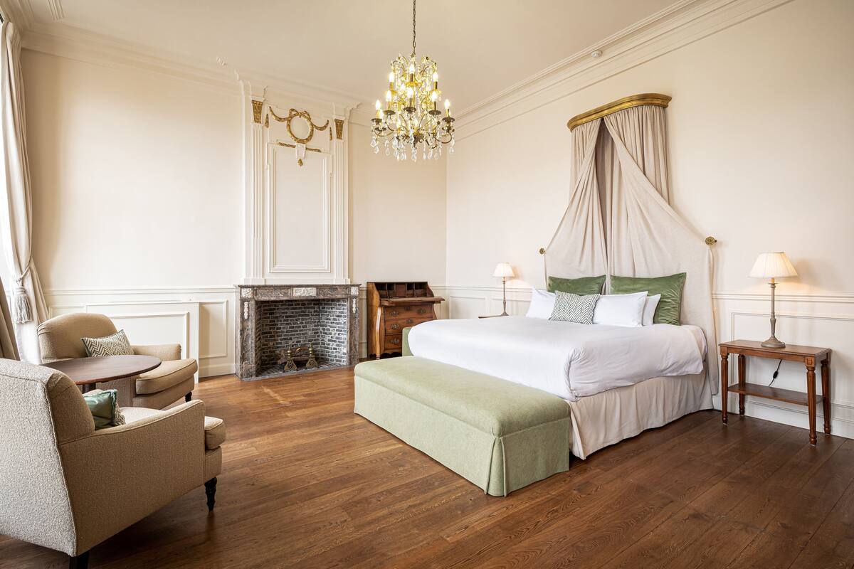 A bedroom with a large bed, a chair, a bench, a fireplace, and a chandelier.