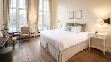 Een hotelkamer met een groot bed, een stoel, een bureau en een nachtkastje. Er zijn twee ramen met gordijnen, een wandlamp en een ingelijst certificaat aan de muur.