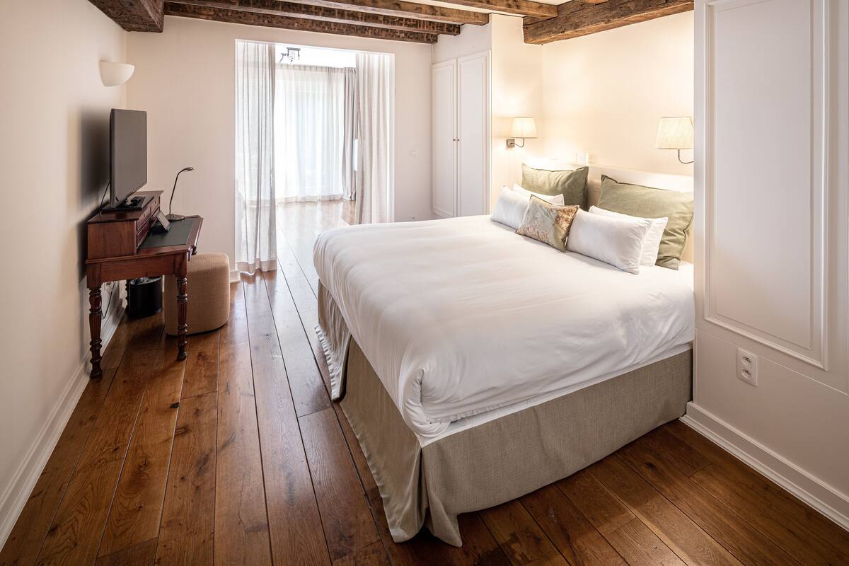 A bedroom with a large bed, wooden flooring, a desk with a television, and a window with curtains.