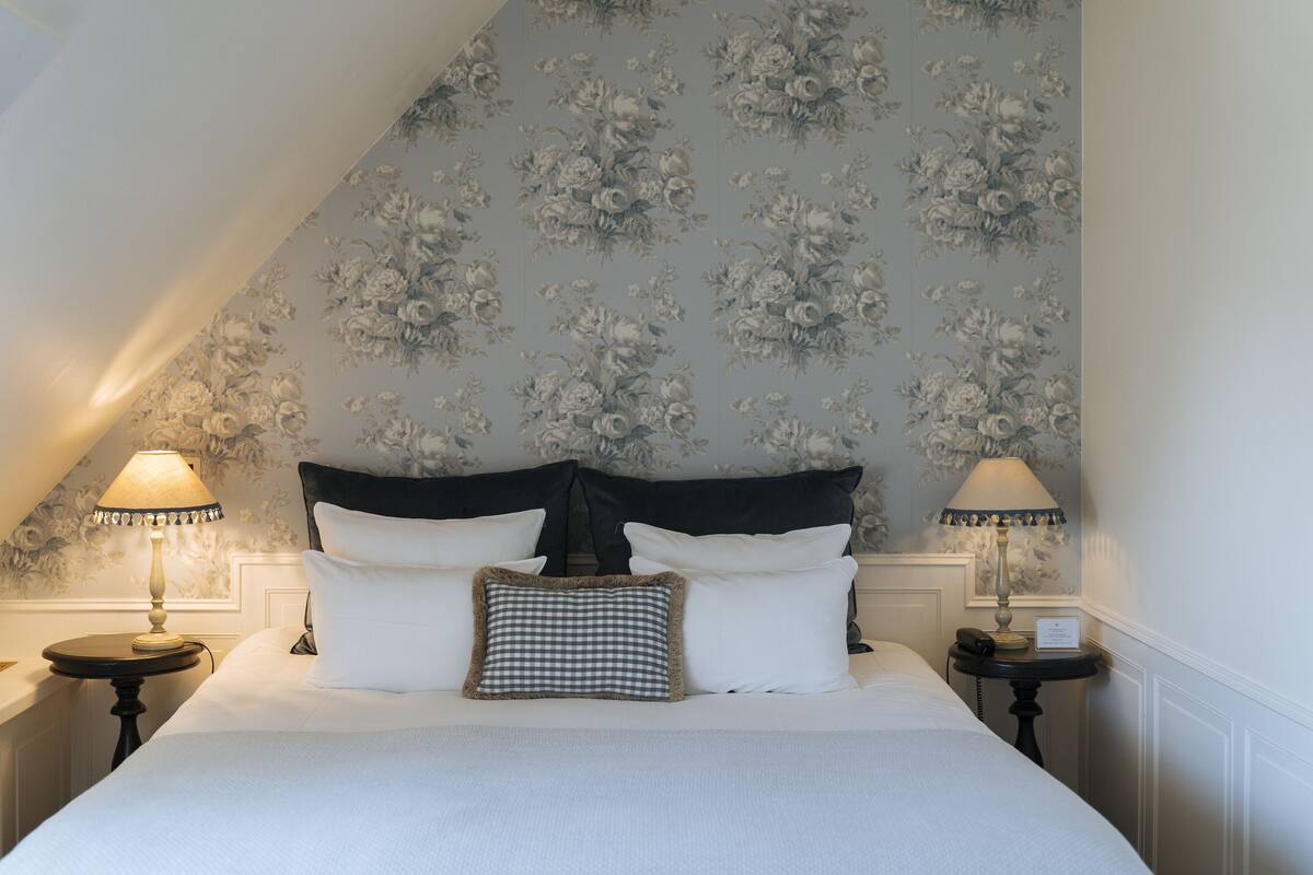 A neatly made bed with white linens and a checkered pillow, flanked by two bedside tables with lamps. The room features a floral wallpaper pattern and a sloped ceiling.