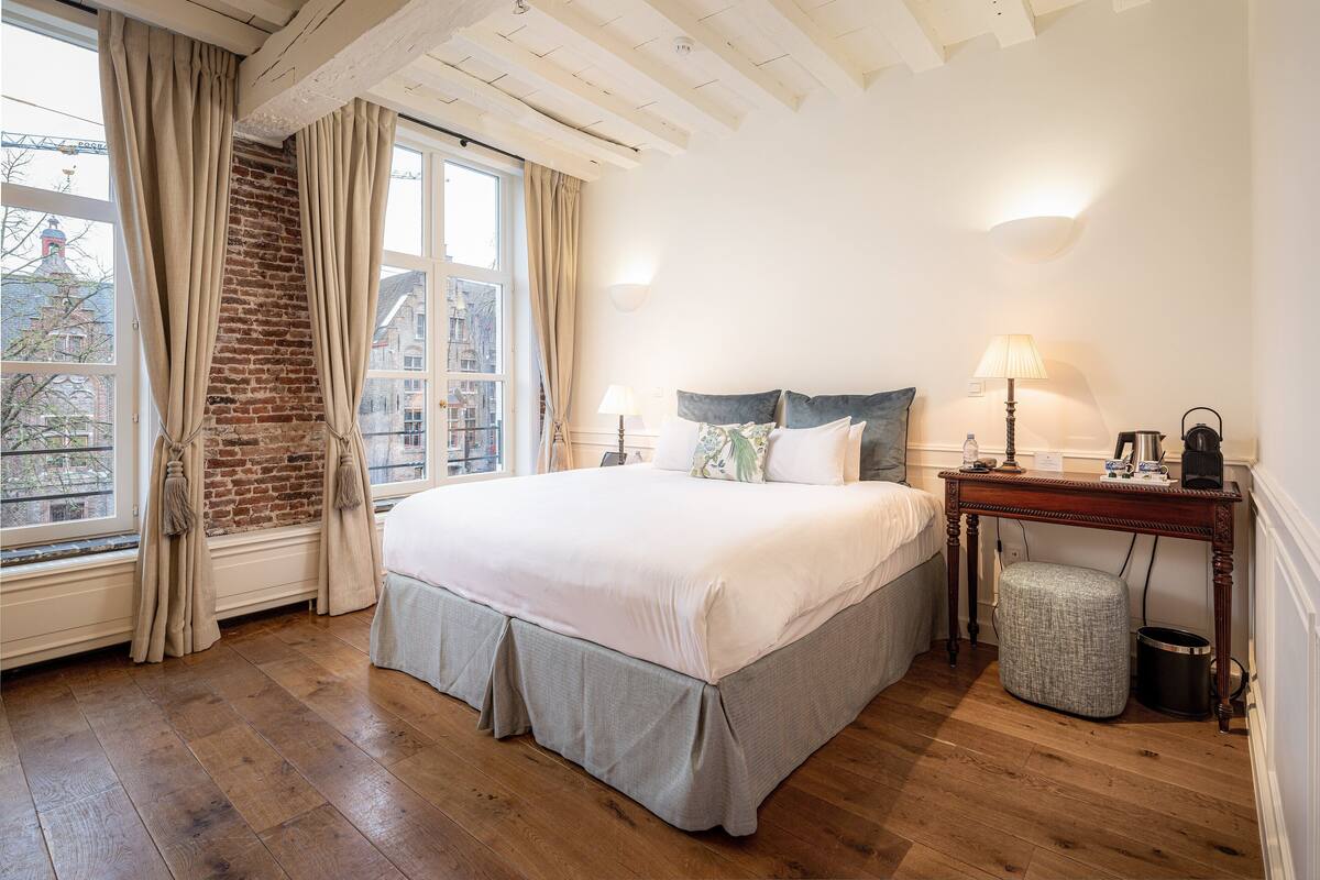 A bedroom with a large bed, a wooden floor, a brick wall, and a window with curtains.