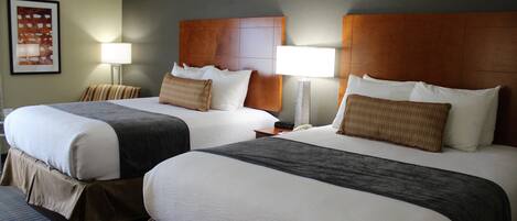 Habitación de hotel con dos camas, cada una con cabecero de madera, ropa de cama blanca y una manta marrón. Hay lámparas de noche y un cuadro en la pared.