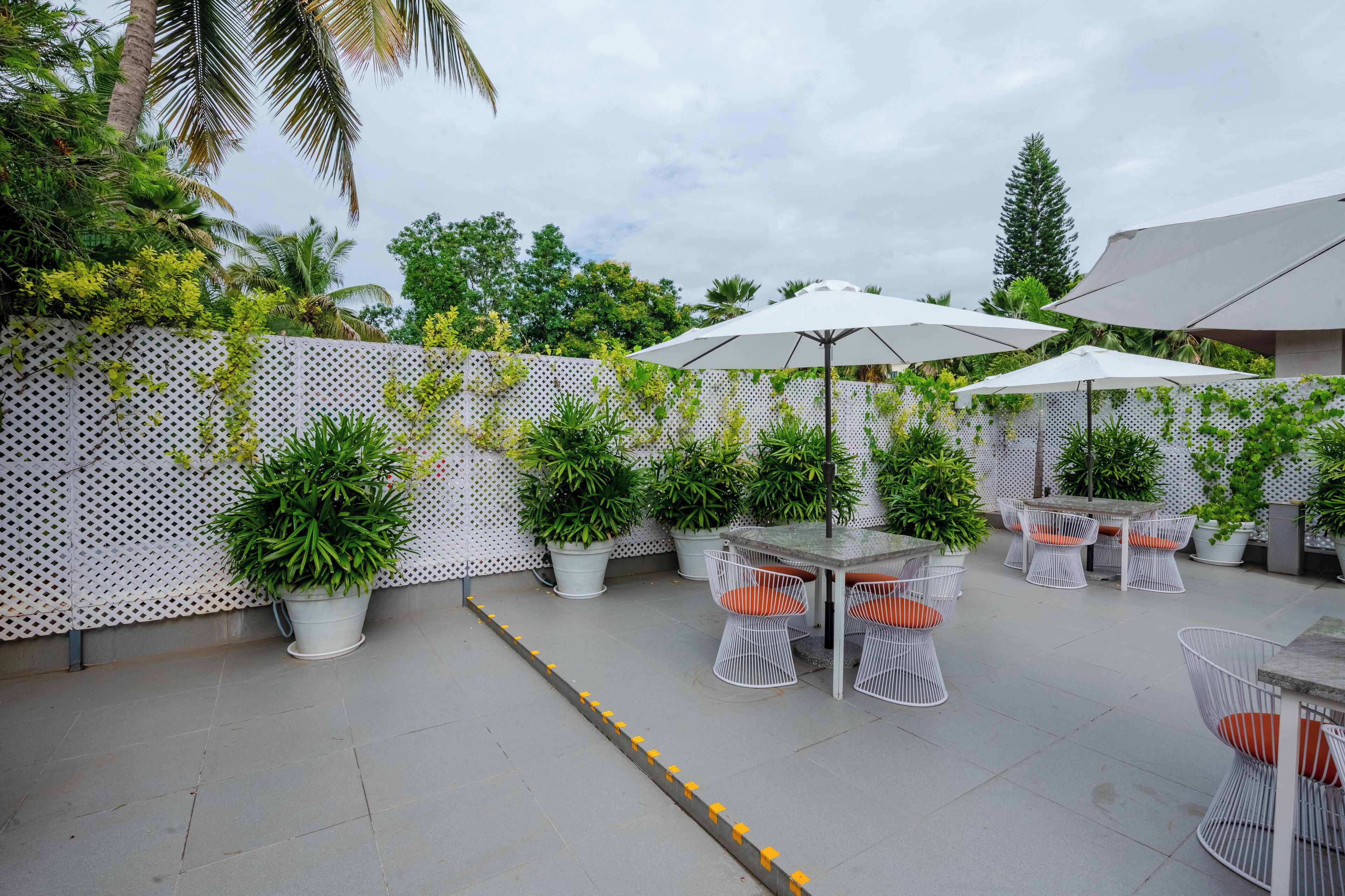 An outdoor patio area with tables and chairs, surrounded by potted plants and a white lattice fence.