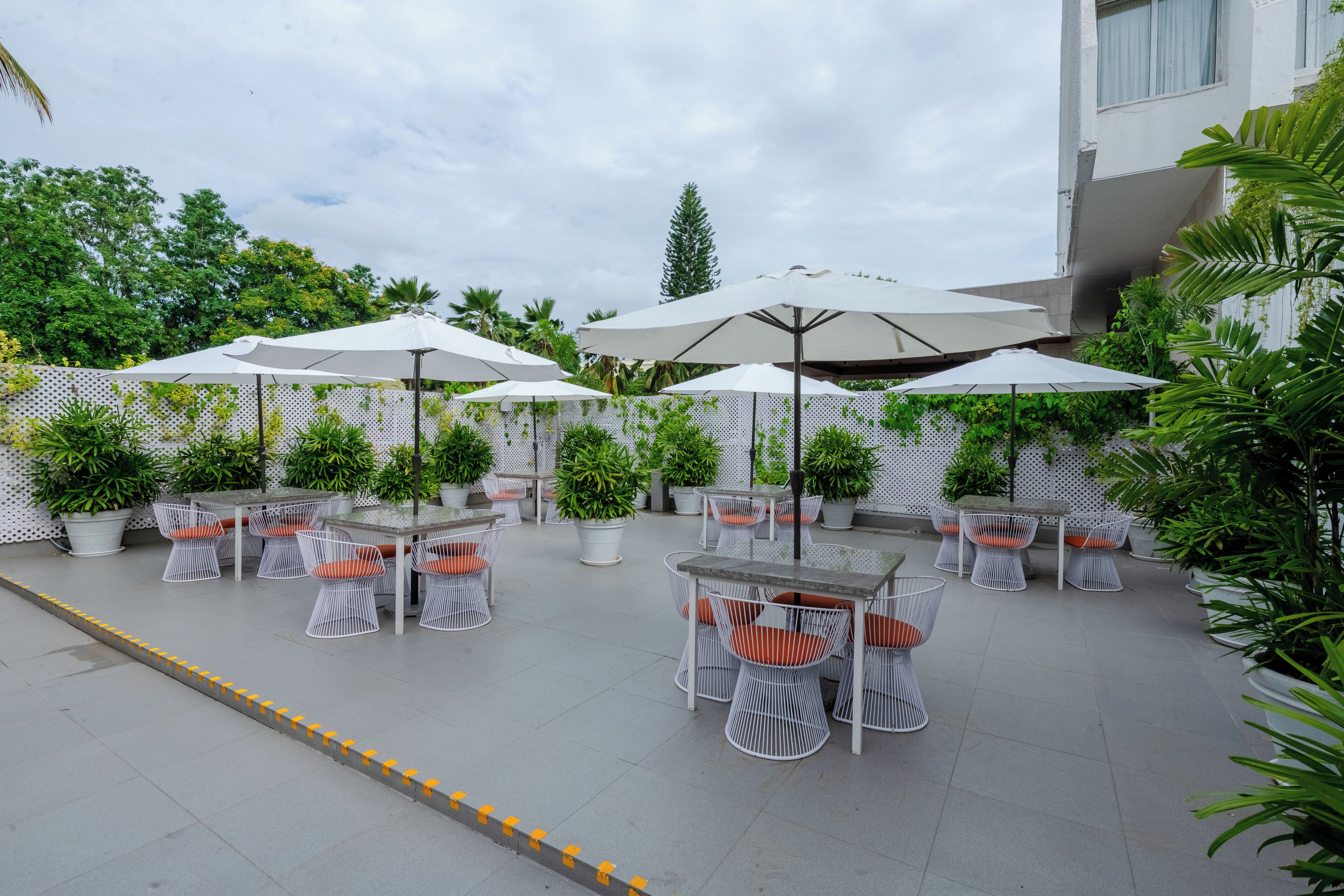 An outdoor patio area with white metal tables and chairs, surrounded by potted plants and umbrellas.