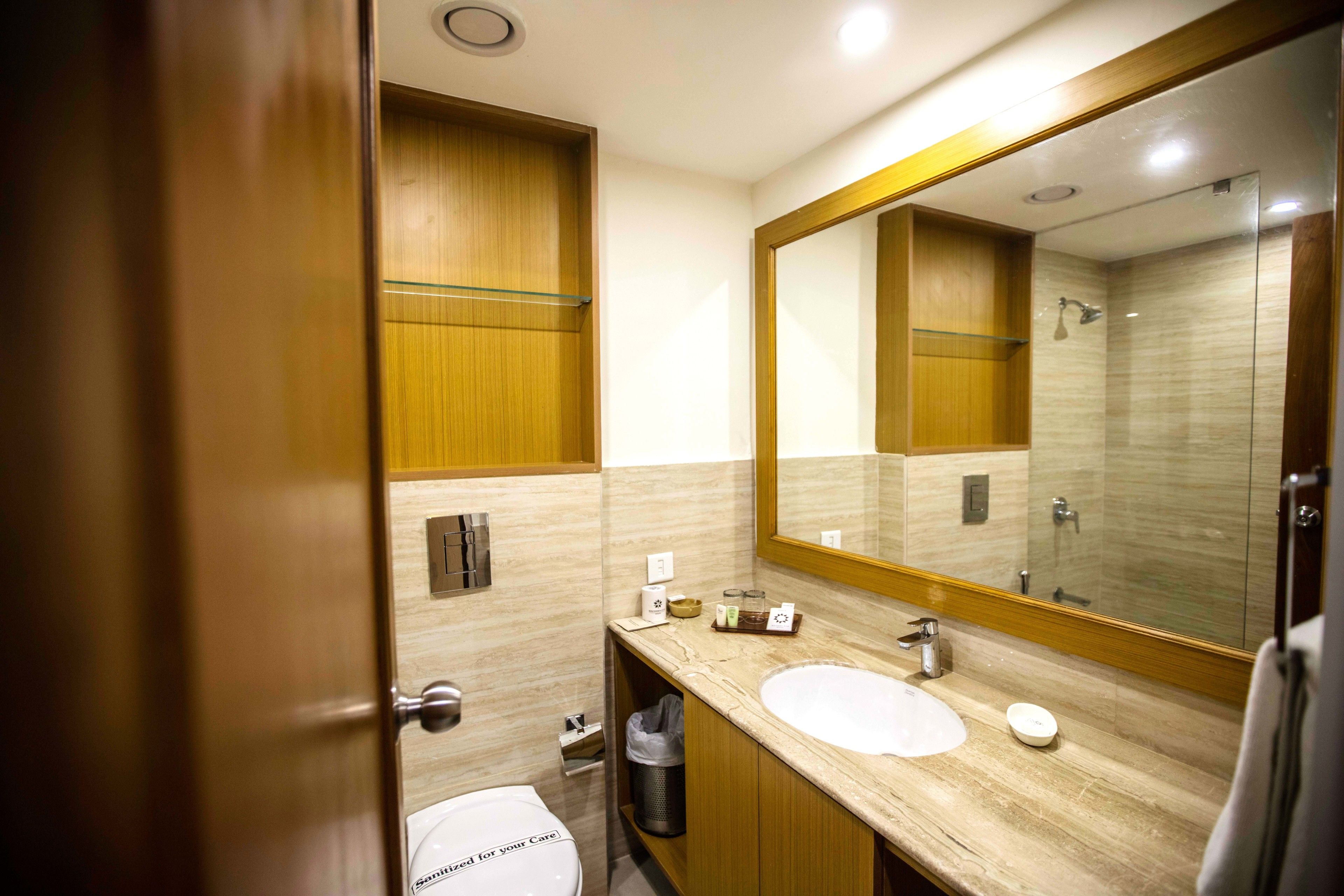 A modern bathroom with a marble countertop, a large mirror, a glass shower enclosure, and a toilet with a "Sanitized for your Care" label.