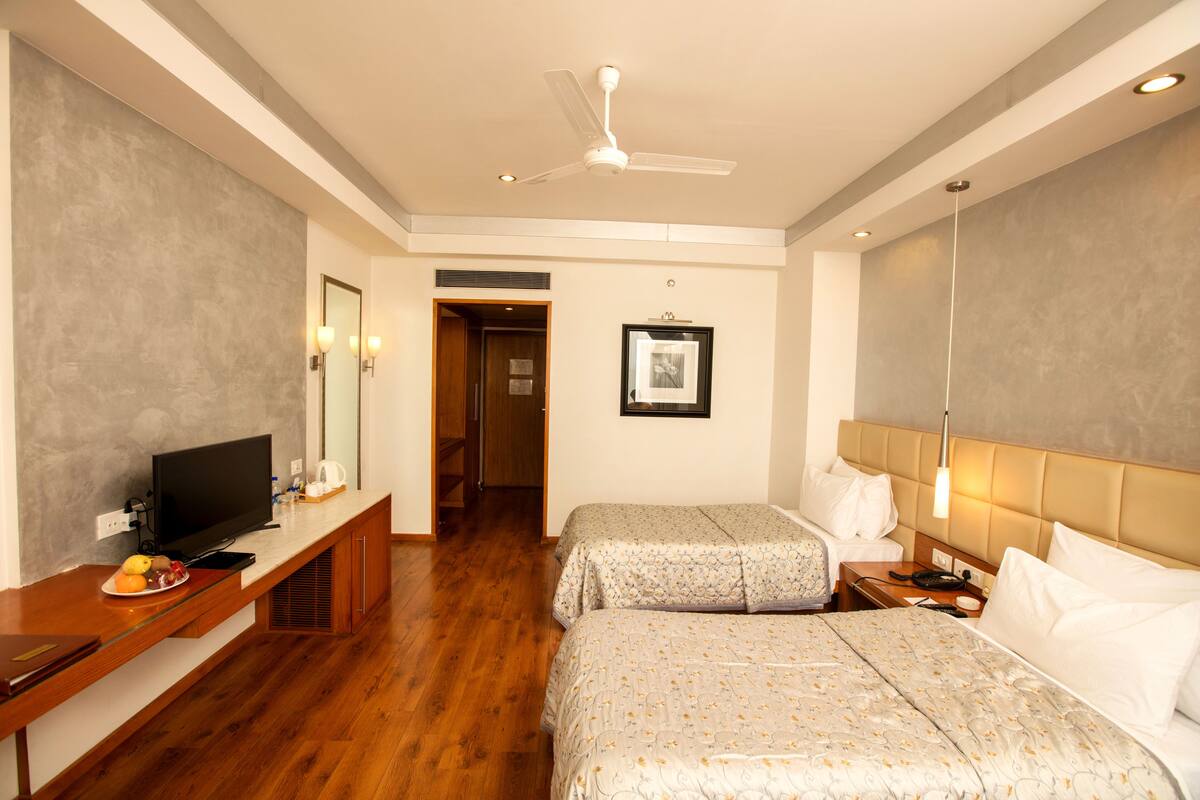 A hotel room with two beds, a television on a wooden console, a ceiling fan, and a framed picture on the wall.