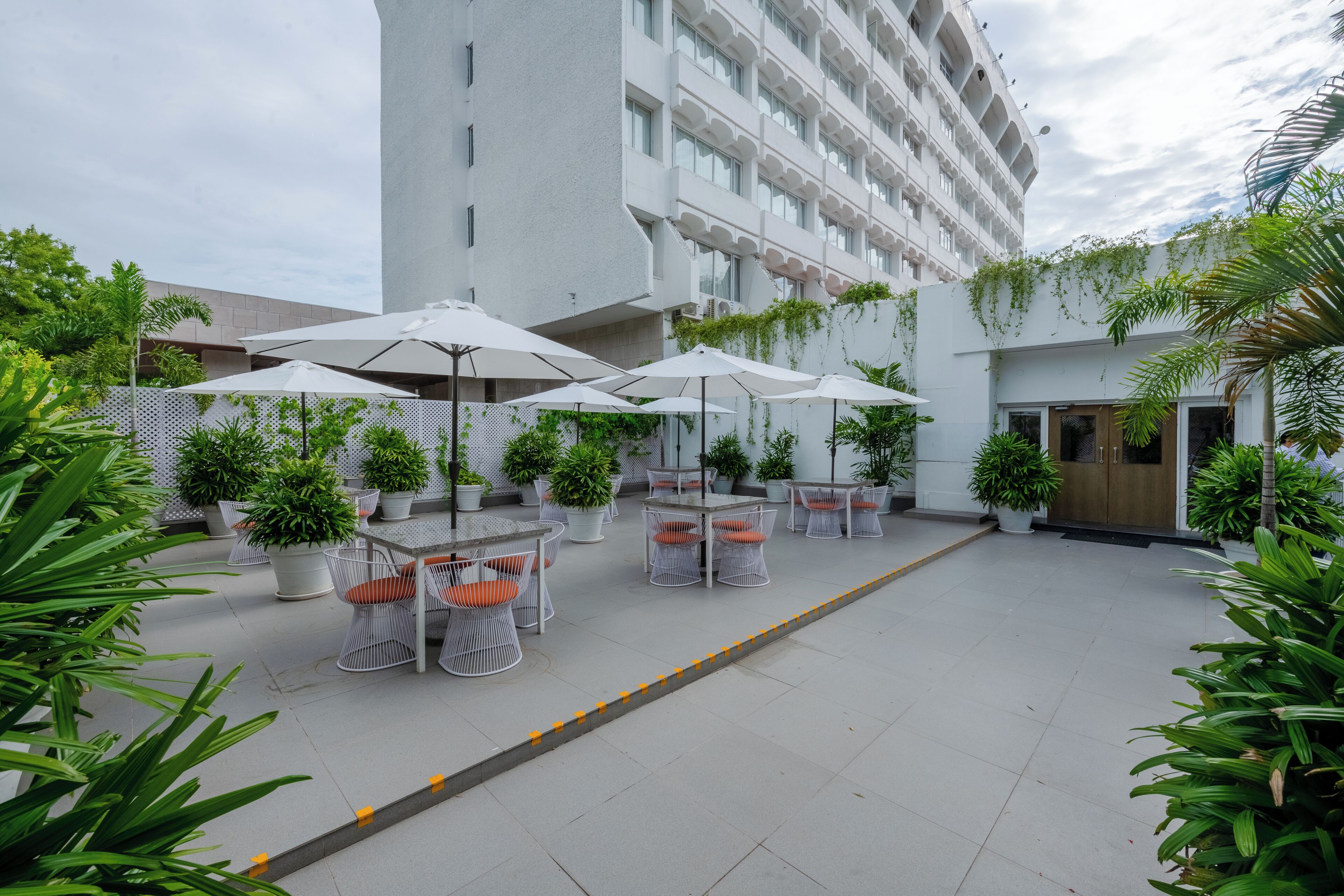 Un patio al aire libre con mesas y sillas, rodeado de plantas en macetas y un edificio grande al fondo.