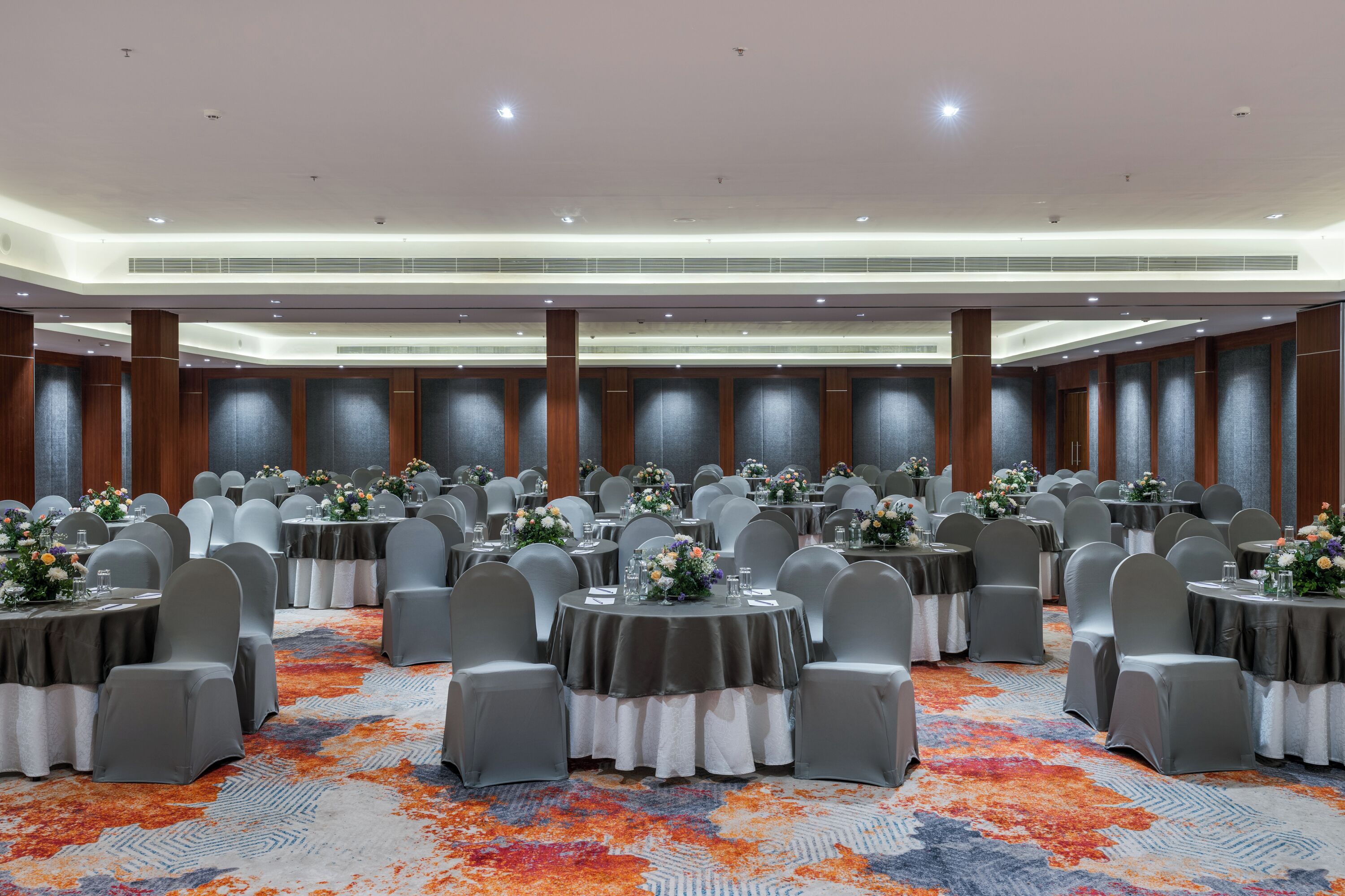 A large banquet hall with round tables, grey chairs, and floral centerpieces.