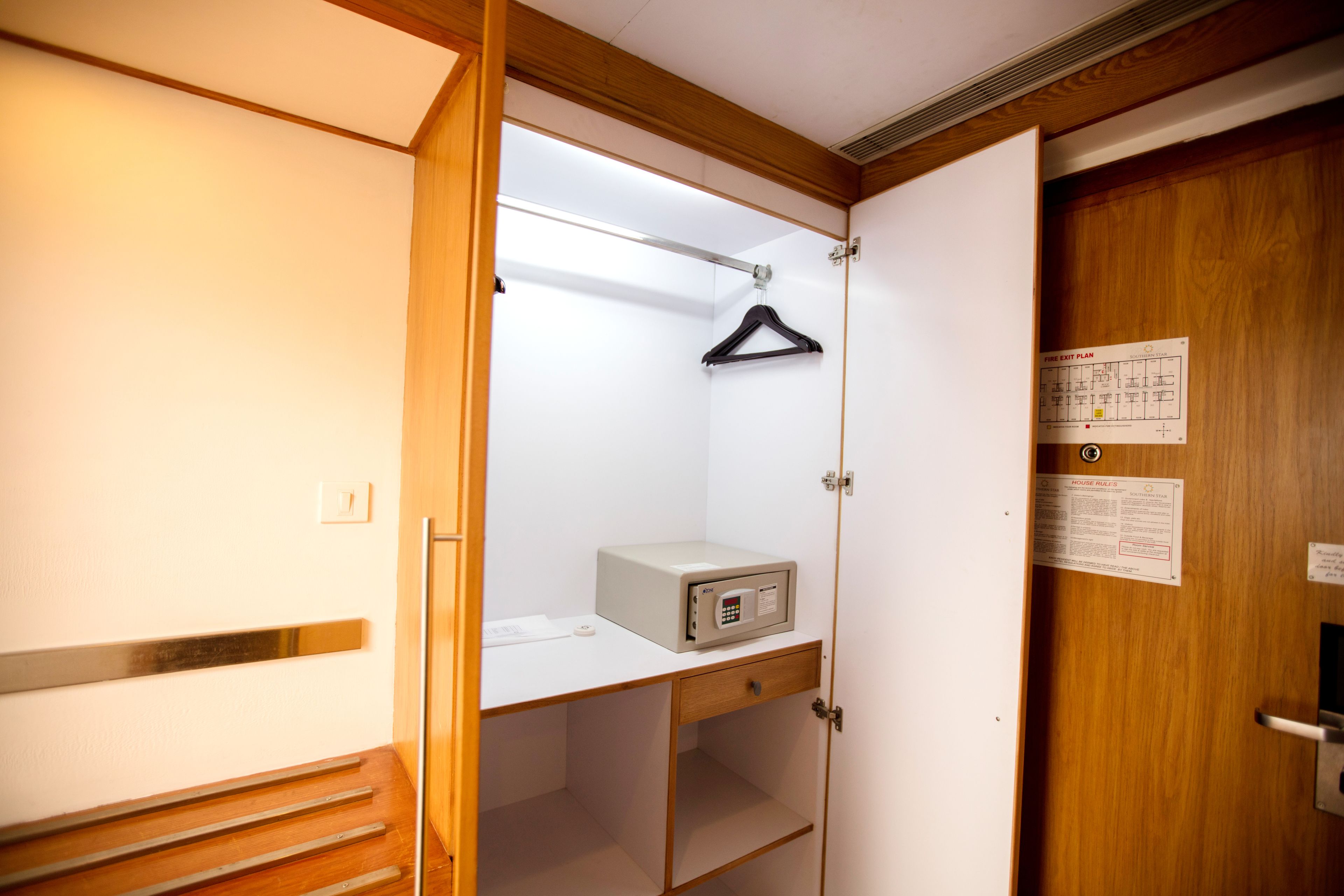 A hotel room with a wooden wardrobe, a desk with a small appliance, and a door with a fire exit plan.