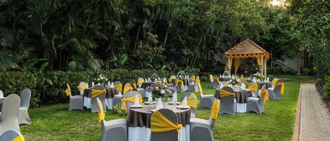 An outdoor event space with round tables and chairs, a gazebo, and lush greenery.