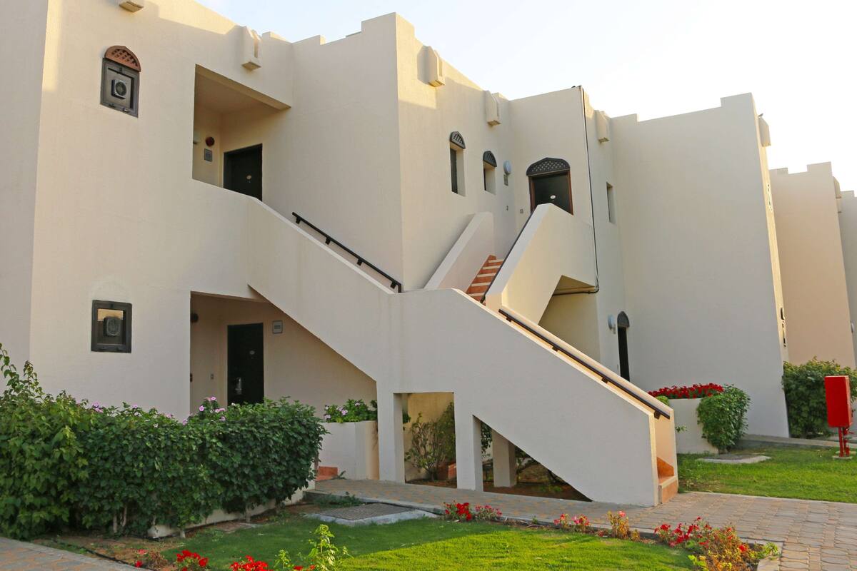 A modern building with a central staircase, multiple balconies, and a well-maintained garden with red flowers.