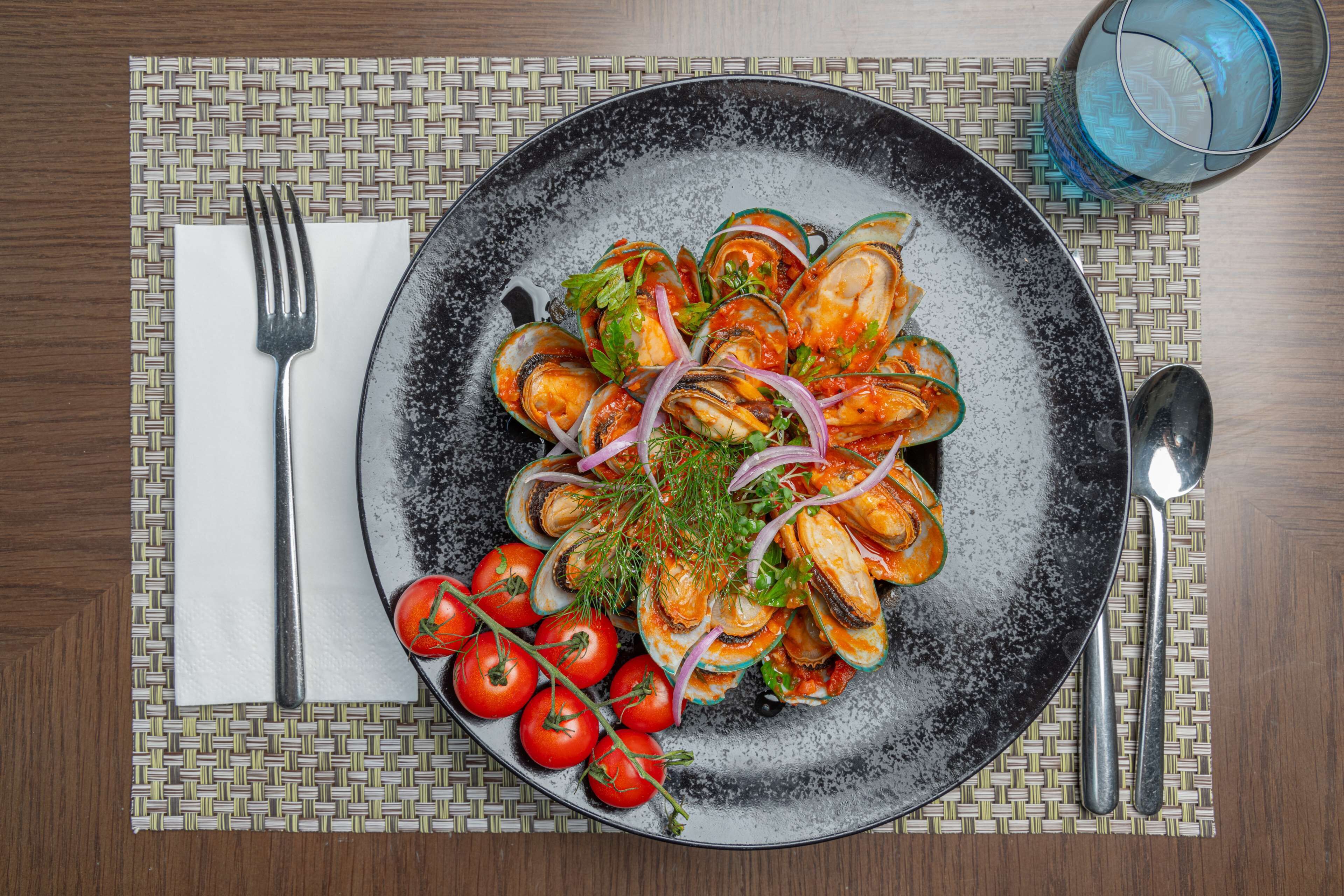 A plate of mussels with tomatoes and herbs, a fork, a spoon, and a glass of water on a table.