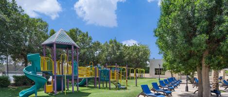 A playground with a slide and climbing structures, surrounded by a lawn and lounge chairs.