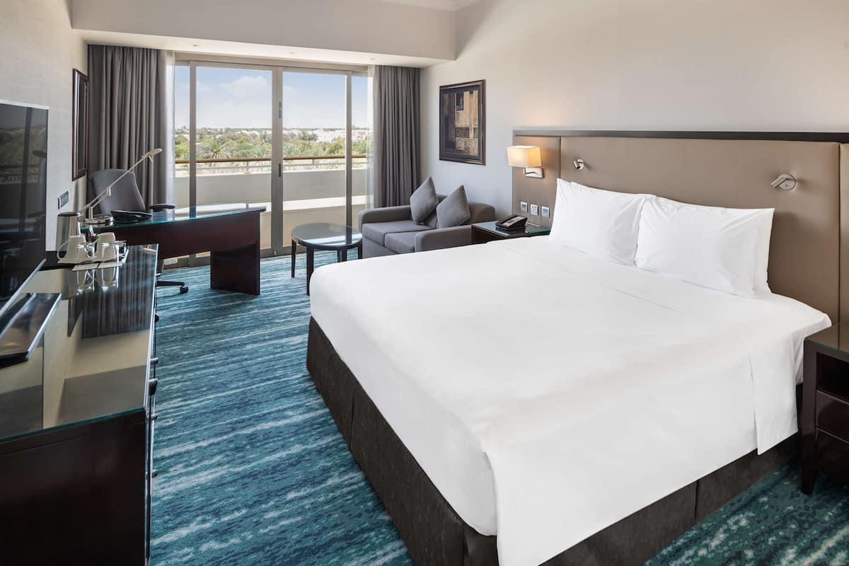 A hotel room with a large bed, a desk with a computer, a chair, a small table, and a view of the outdoors.