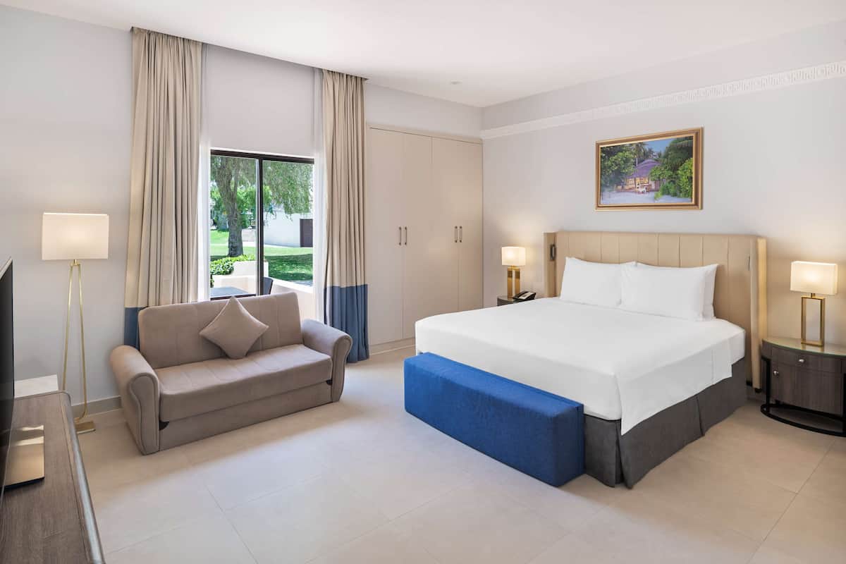 A modern hotel room with a large bed, a sofa, a desk, and a view of the outdoors.