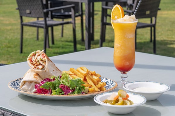 Una comida que incluye un wrap, papas fritas y una bebida sobre una mesa al aire libre.