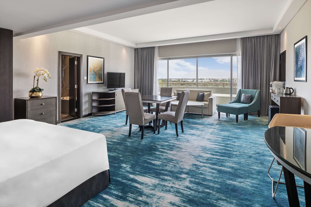 A hotel room with a large bed, a dining area with chairs, a TV, and a view of the city.