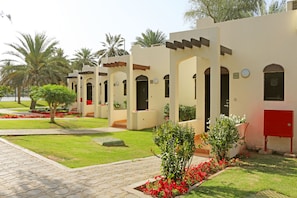 A row of modern, beige-colored buildings with arched doorways and windows, surrounded by well-maintained lawns and palm trees.
