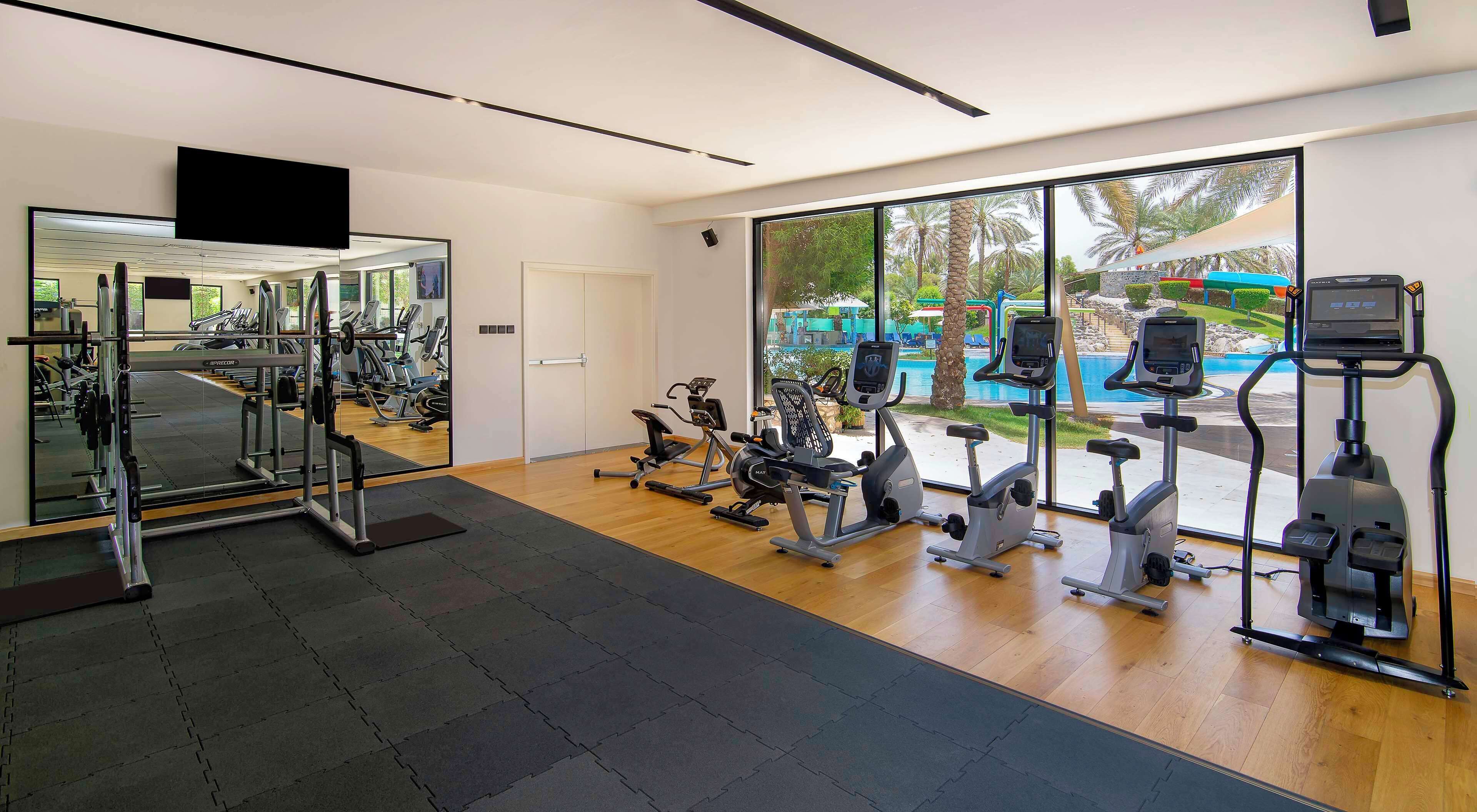 A modern gym with cardio machines, weightlifting equipment, and large windows offering a view of an outdoor pool area.