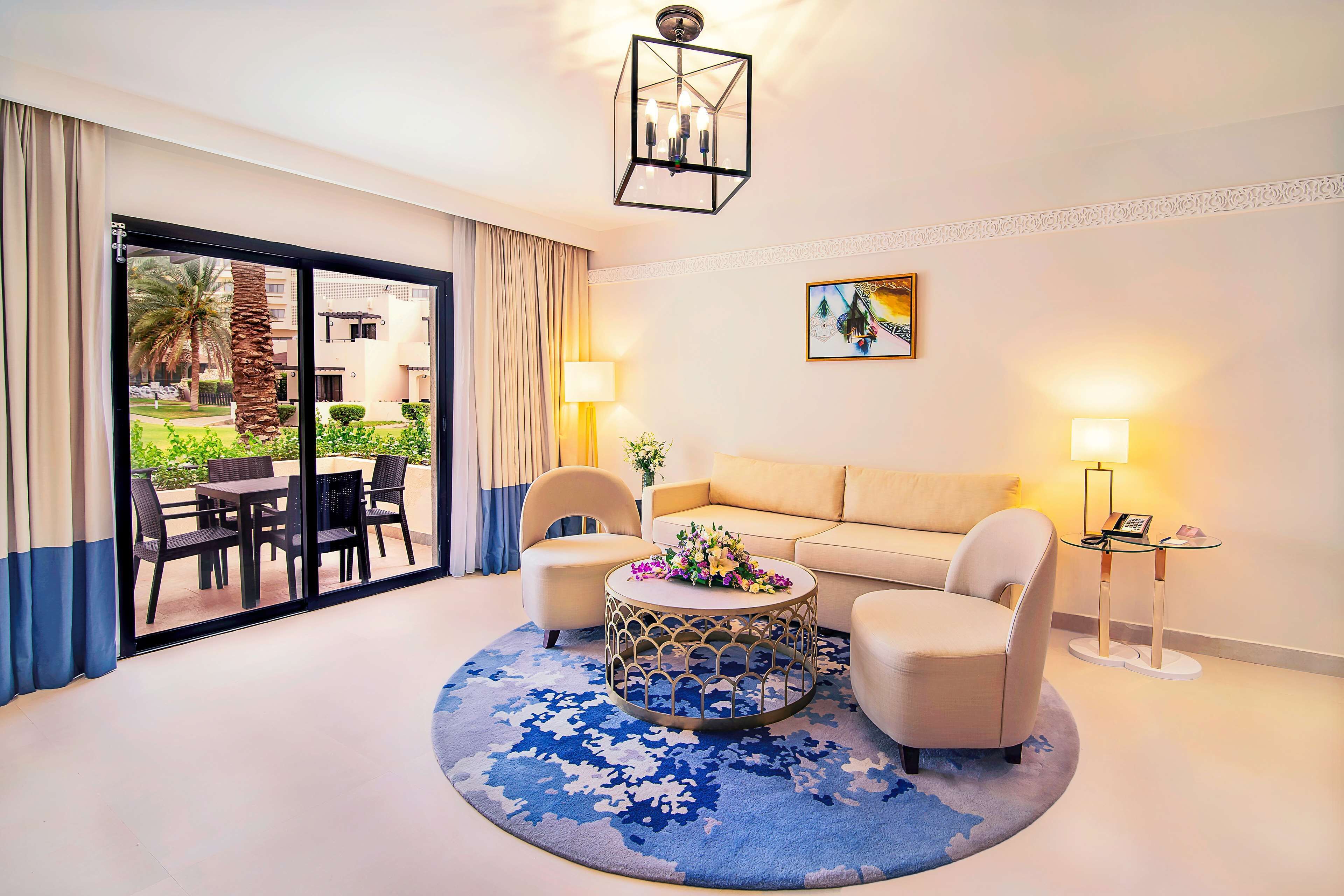 Een moderne hotelkamer met een ronde salontafel, twee fauteuils, een bank, een groot raam met gordijnen, een kroonluchter en een schilderij aan de muur.