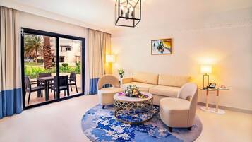 Een moderne hotelkamer met een ronde salontafel, twee fauteuils, een bank, een groot raam met gordijnen, een kroonluchter en een schilderij aan de muur.