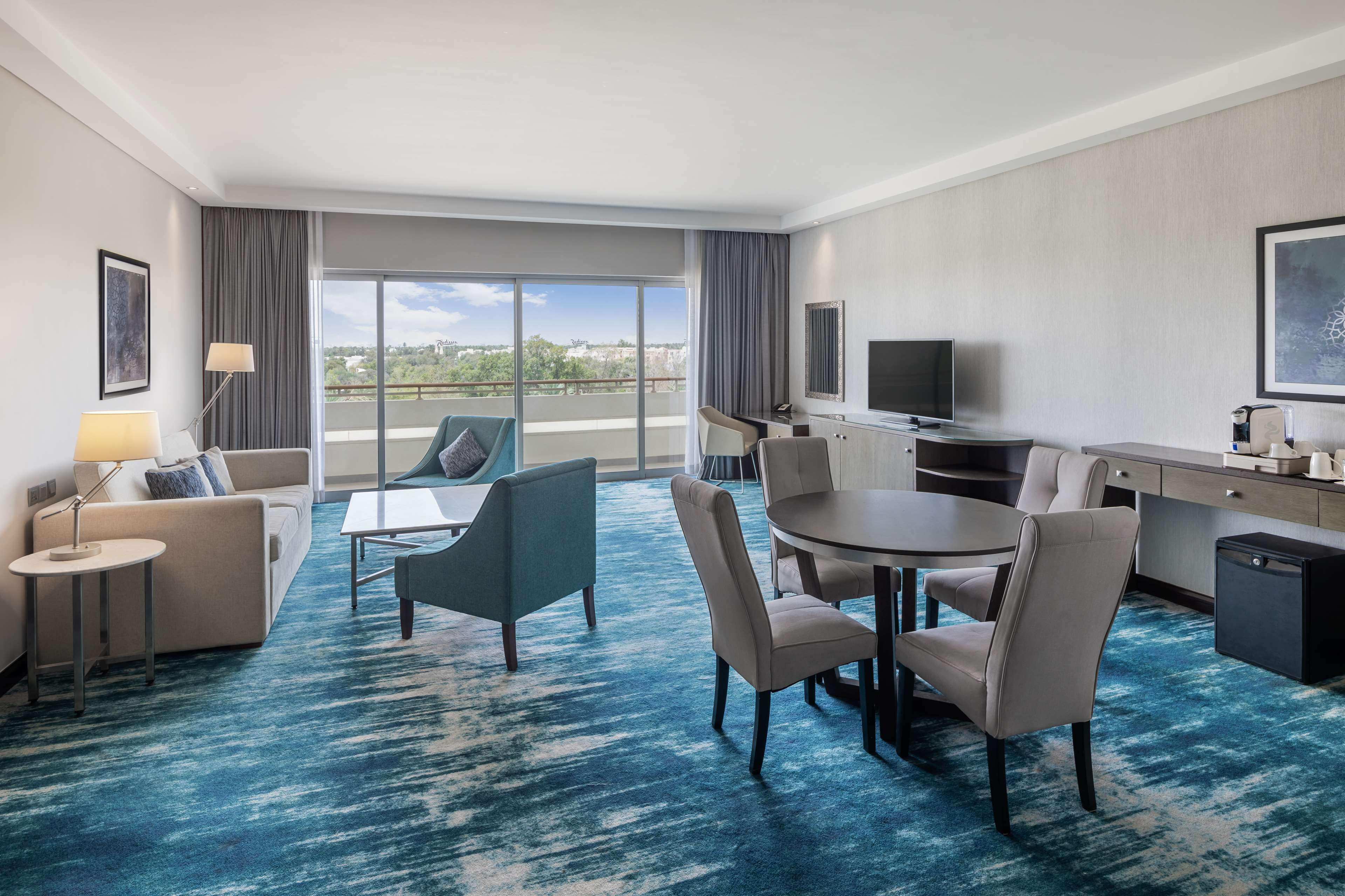 A hotel room with a large window, a dining area with a round table and chairs, a sofa, a TV, and a small kitchen area.