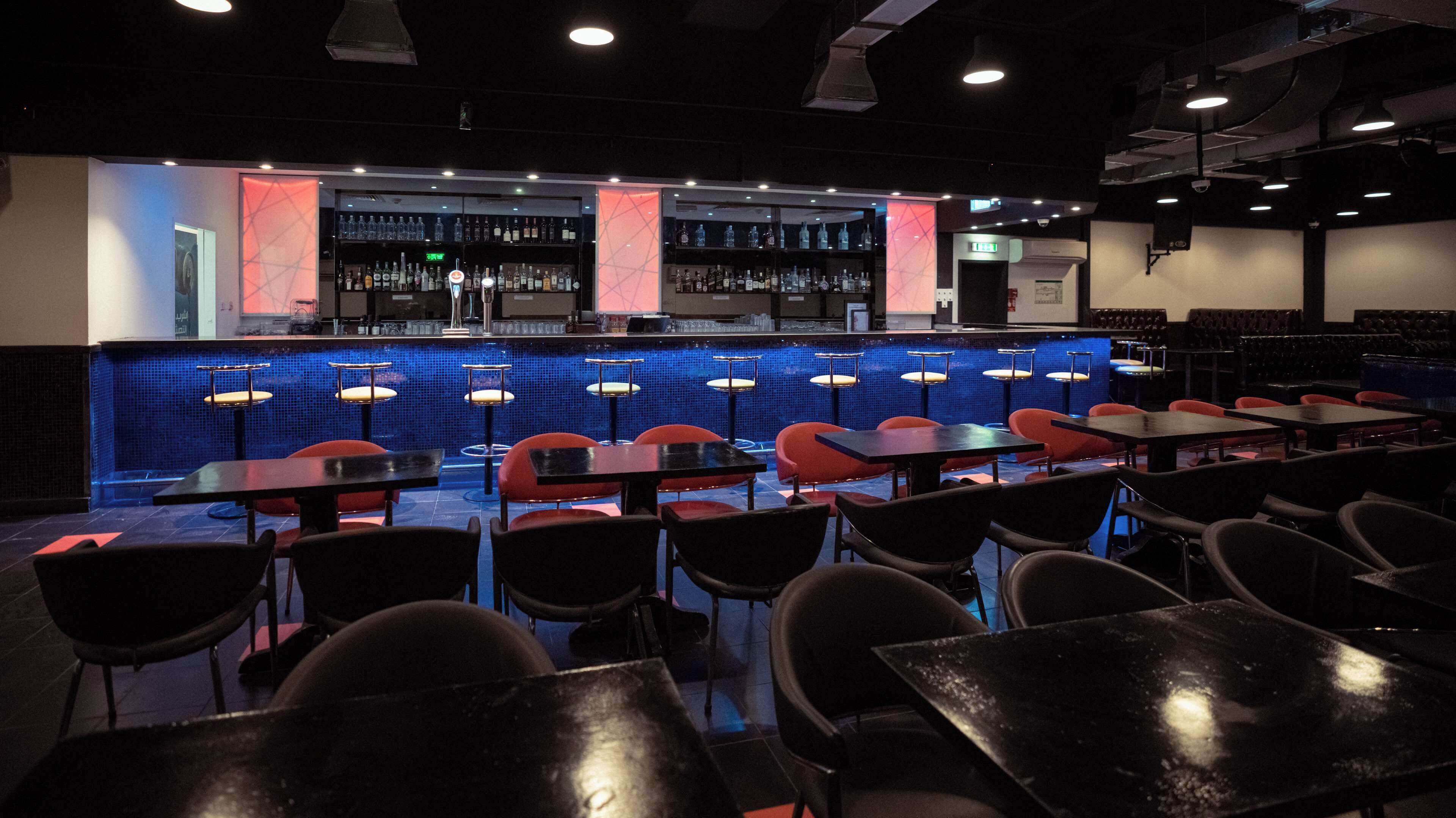 Una zona de bar con mostrador de azulejos azules, sillas rojas y mesas negras.