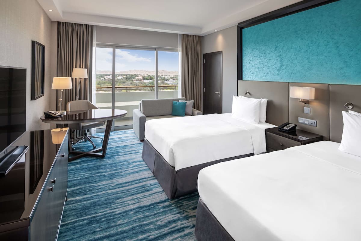 A hotel room with two beds, a desk with a chair, a TV, and a large window with a view.