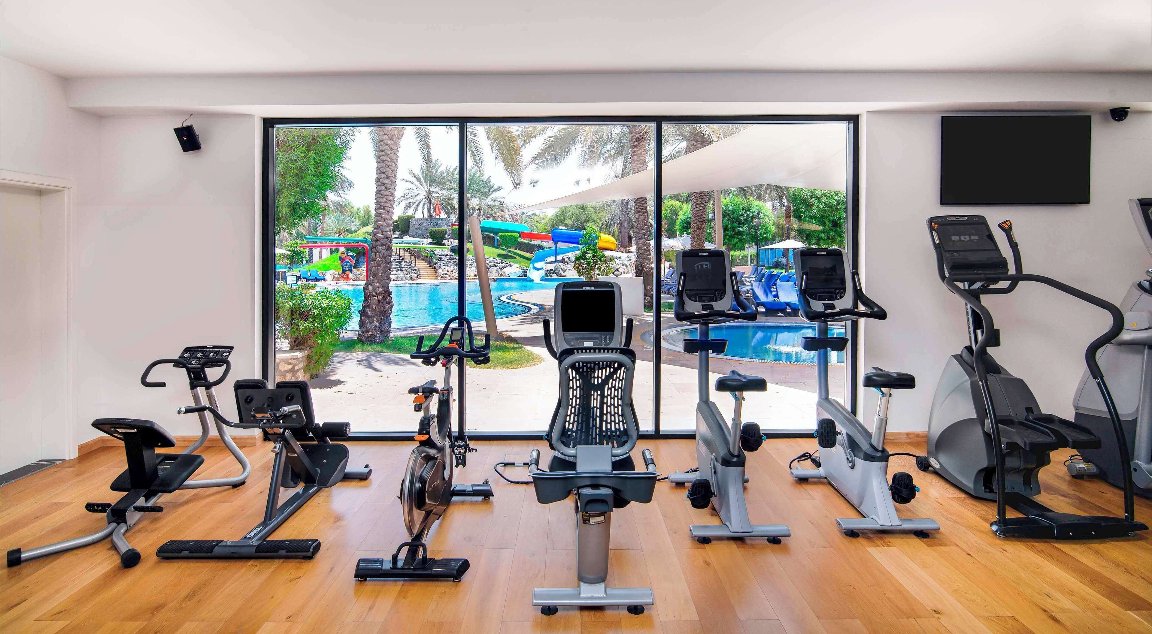 A gym with various exercise machines, large windows with a view of a pool and outdoor area.