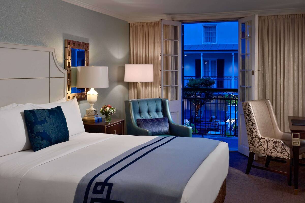 Ein Hotelzimmer mit einem Bett, zwei Stühlen, einem Nachttisch, einer Lampe und Blick ins Freie.