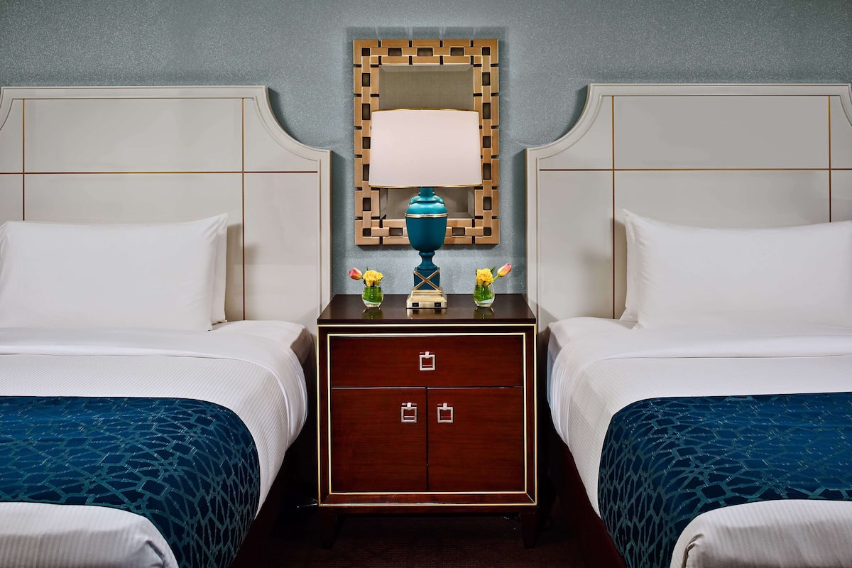 Ein Hotelzimmer mit zwei Betten, einem Nachttisch mit Lampe und Blumenvase sowie einem dekorativen Spiegel.