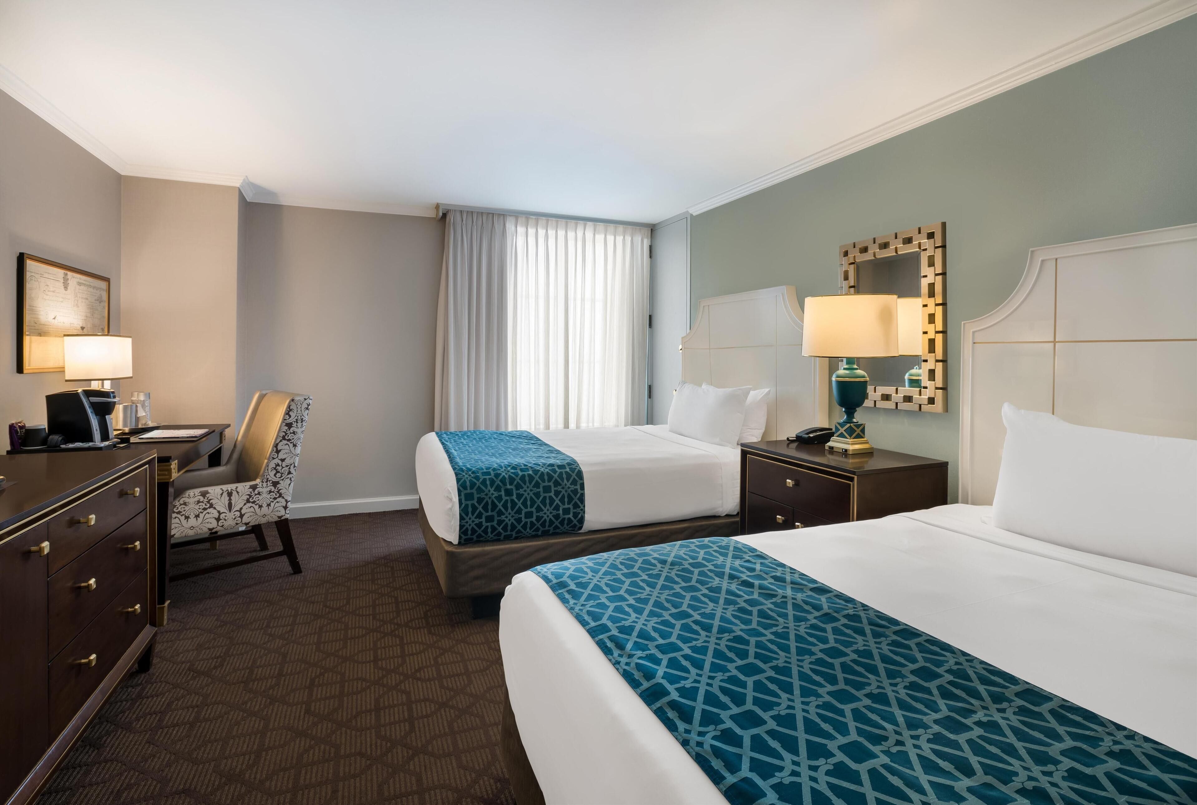 A hotel room with two beds, a desk with a chair, a dresser, and a large window with curtains.