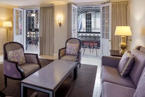 A hotel room with a sofa, two armchairs, a coffee table, a lamp, and a view of a balcony with outdoor furniture.