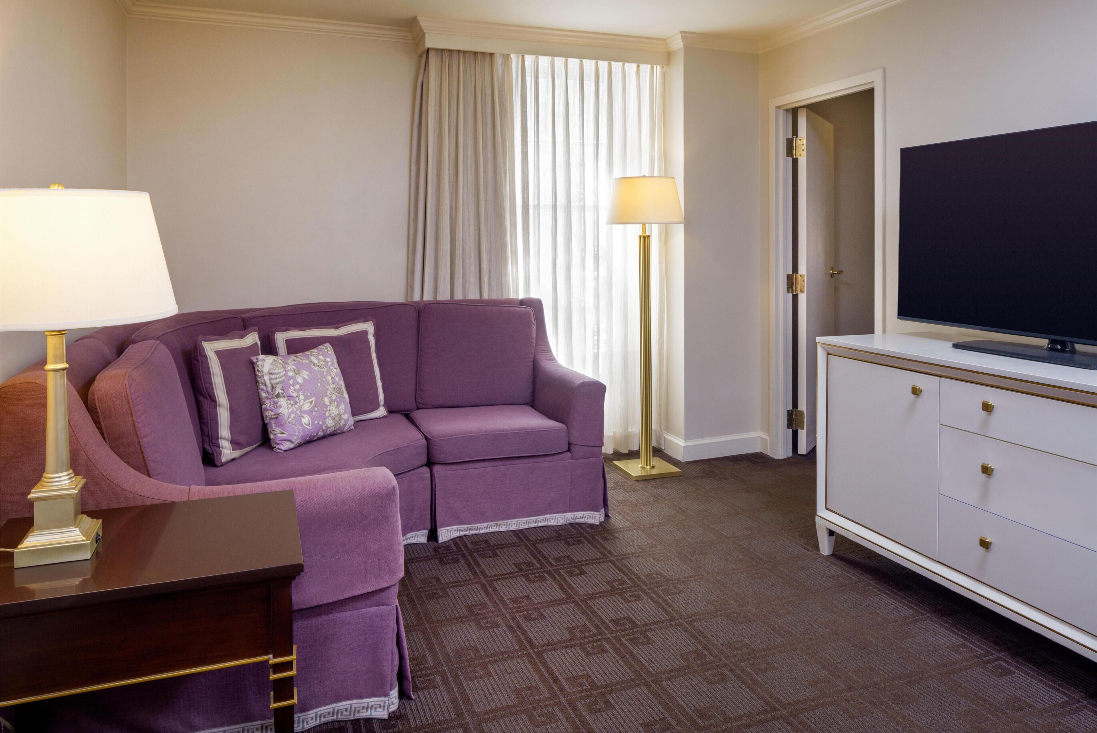 A hotel room with a purple sofa, a TV on a cabinet, a lamp, and a patterned carpet.