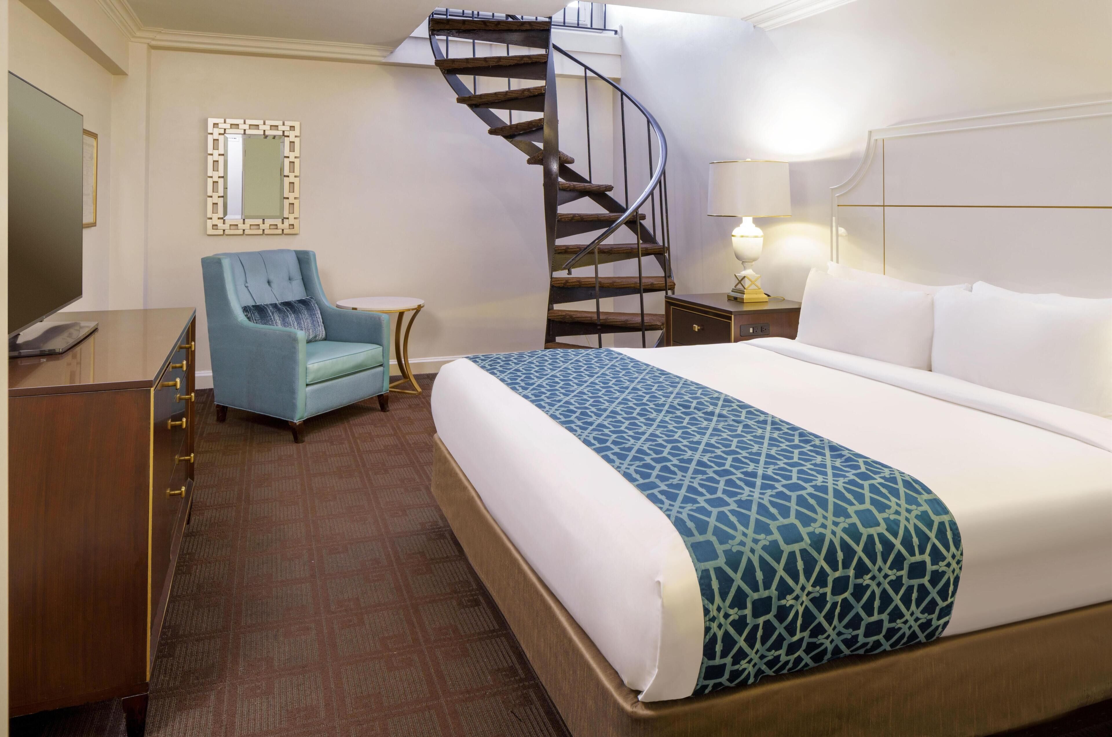Habitación de hotel con cama, una silla, escritorio, televisor, lámpara y una escalera en espiral.