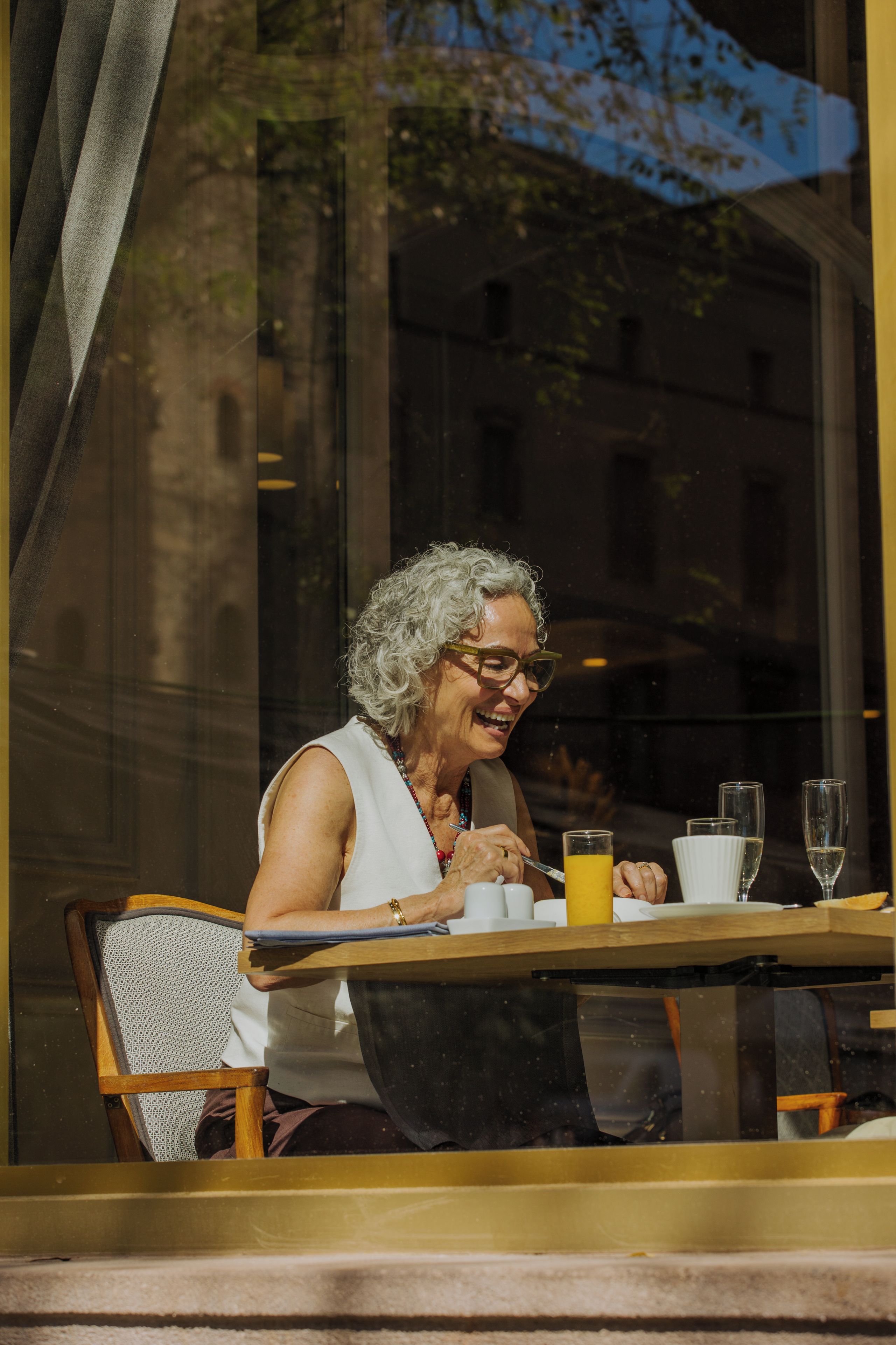 An elderly person seated at a table with a window behind them, a cup, and a glass of orange juice.