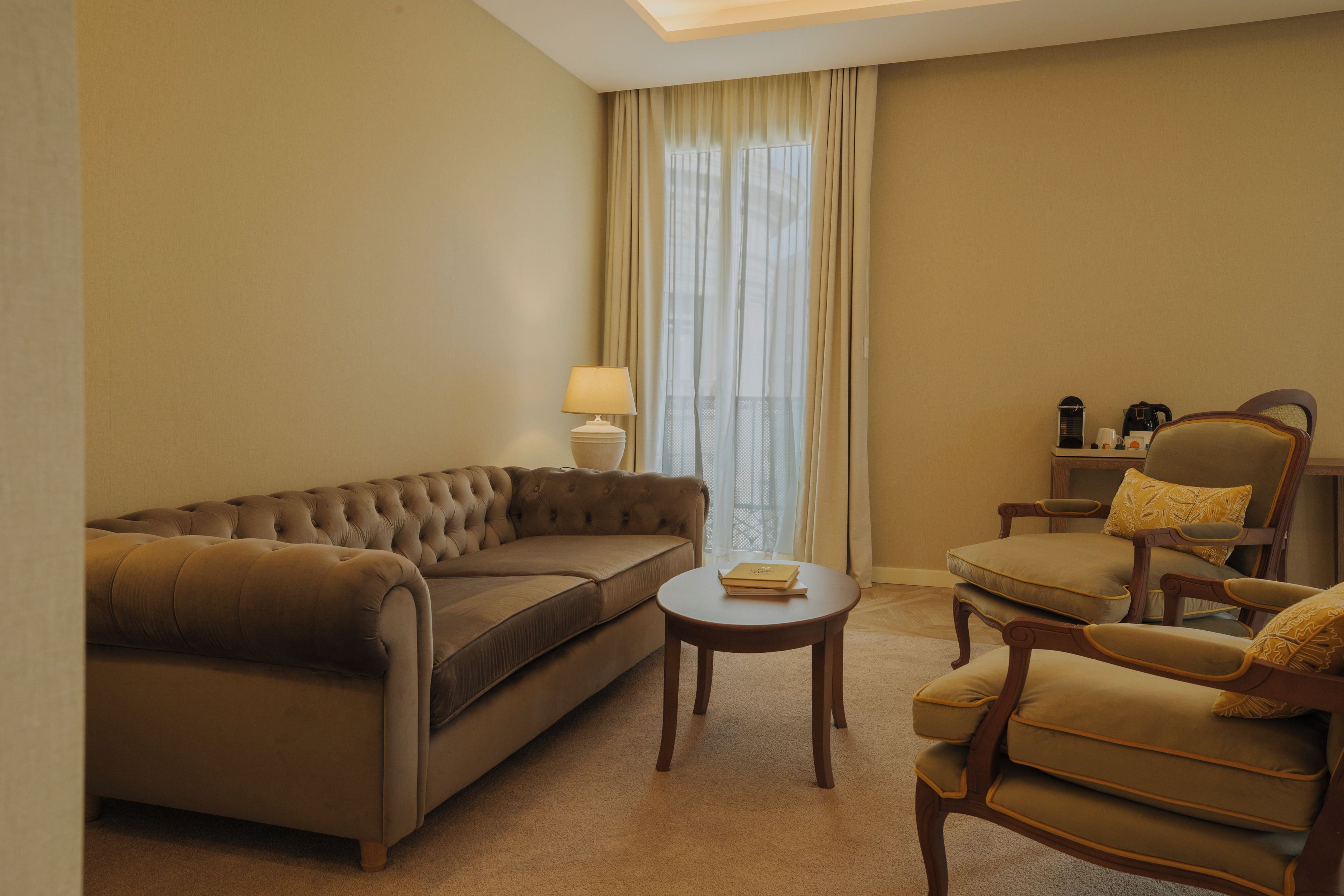 A hotel room with a brown sofa, a wooden armchair, a small round table, and a lamp.