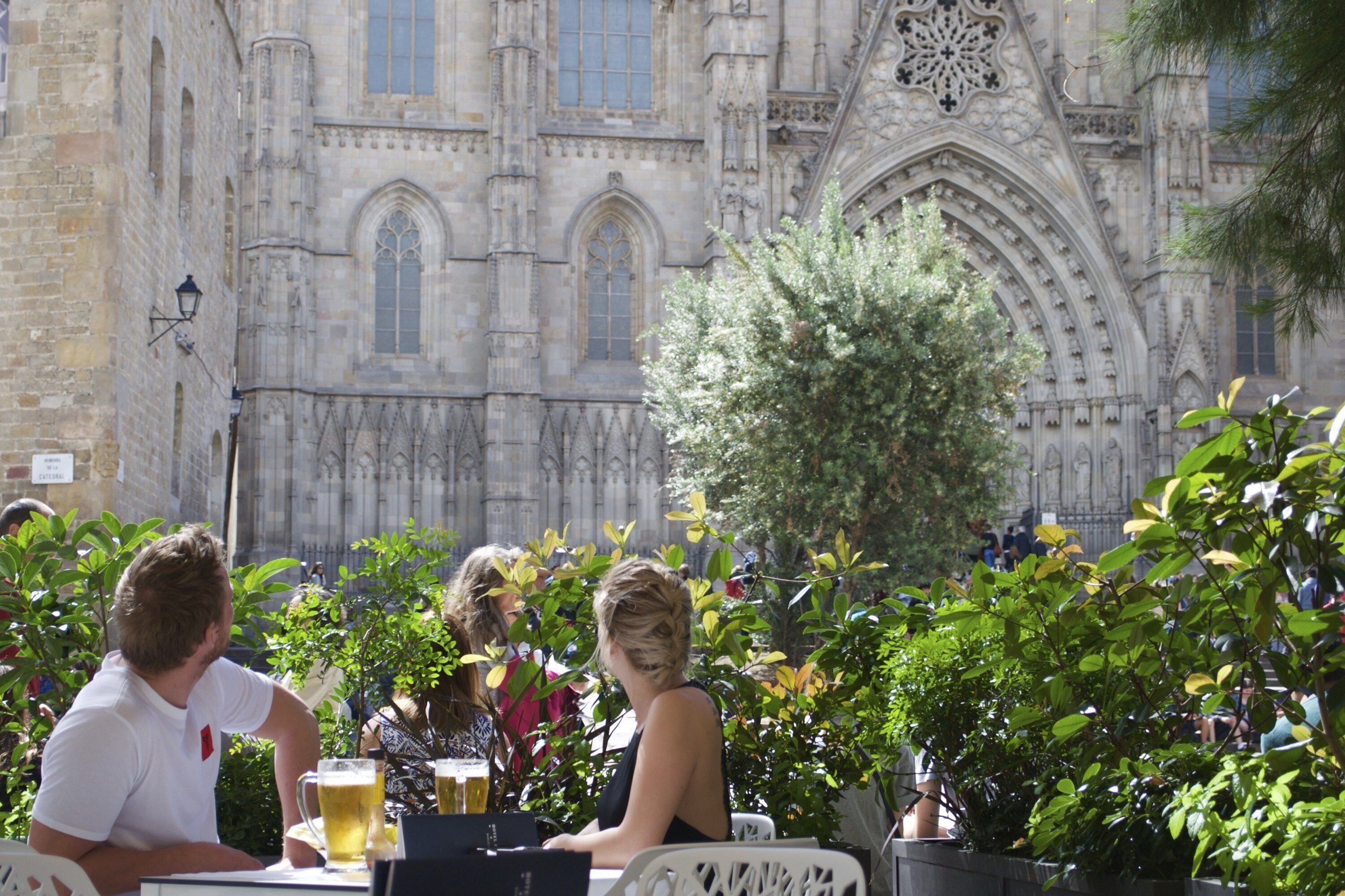 An outdoor café with tables and chairs, a large ornate building in the background, and greenery in the foreground.