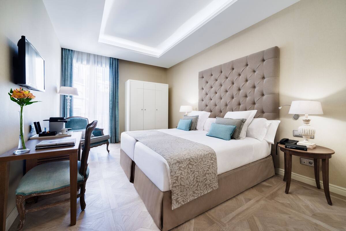 Quarto de hotel moderno com uma cama grande, uma mesa com cadeira, televisão na parede e uma mesinha de cabeceira com abajur.
