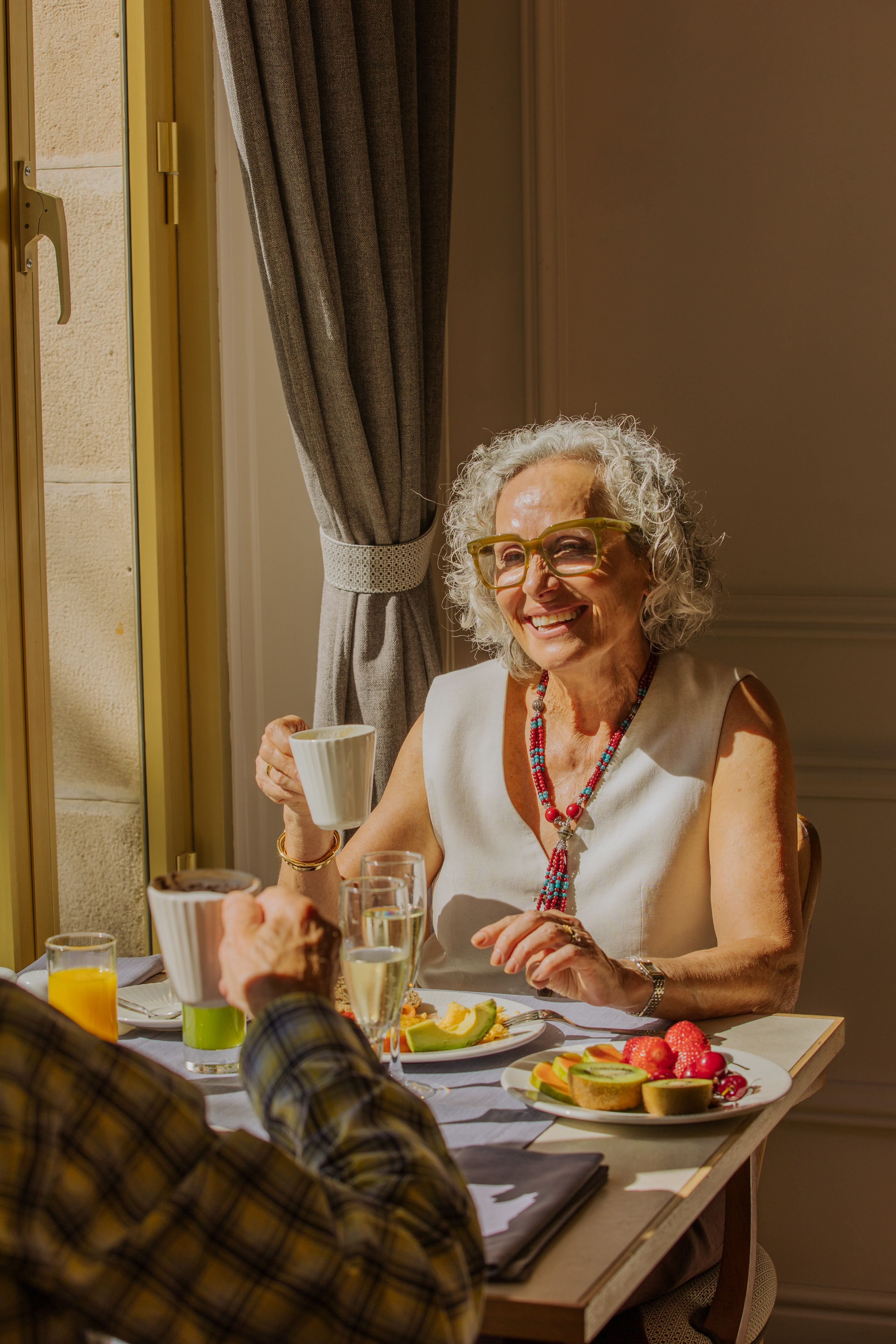 Una persona mayor sentada a una mesa con comida, sosteniendo una taza.