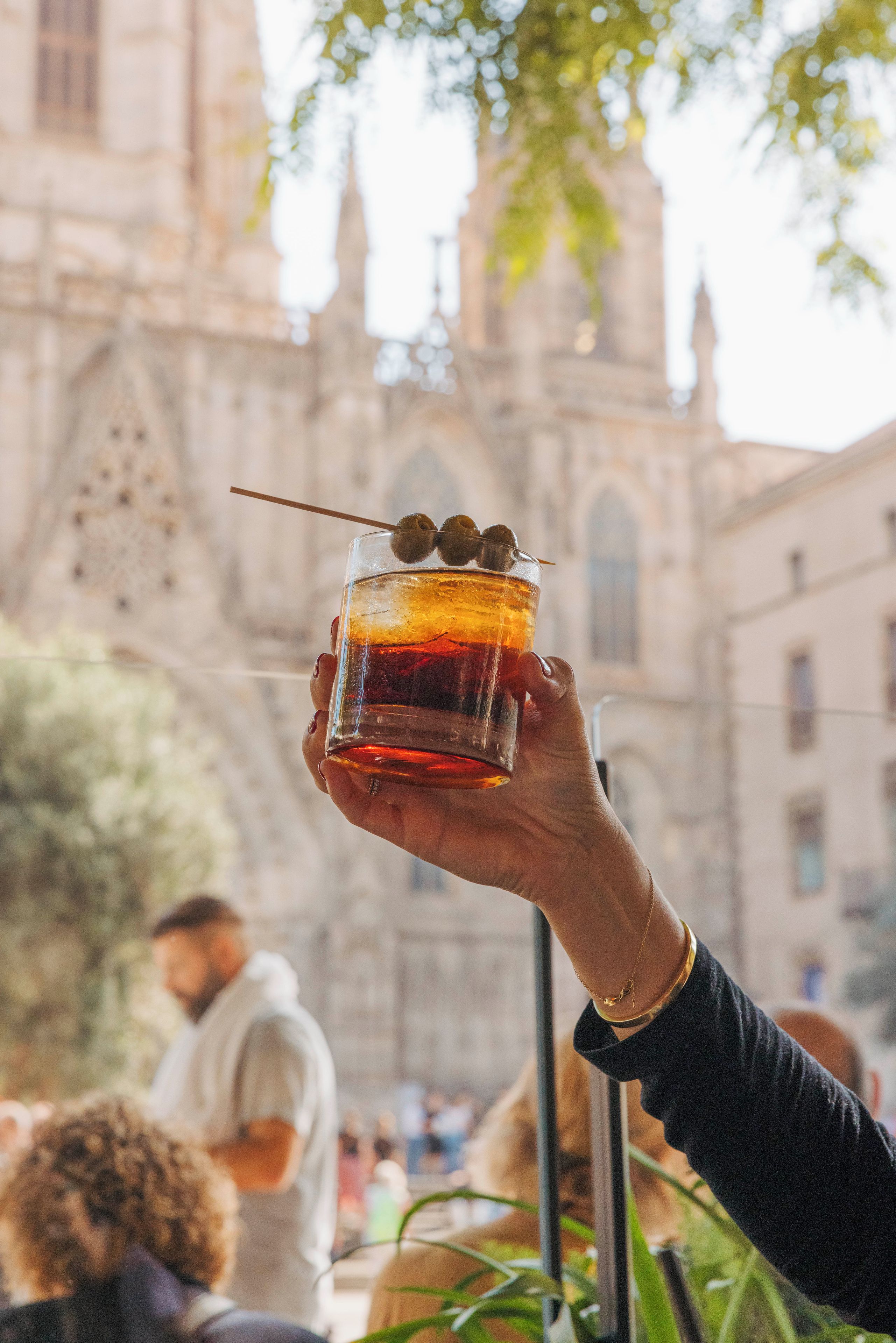 A hand holding a cocktail with olives, set against a blurred background of a building with arched windows and a bell tower.